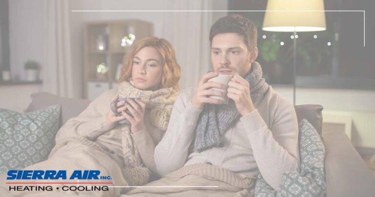 Image: A Couple Sits On The Couch, Bundled Up With Warm Beverages, Cover Image For Why Does My Furnace Fail To Blow Hot Air?